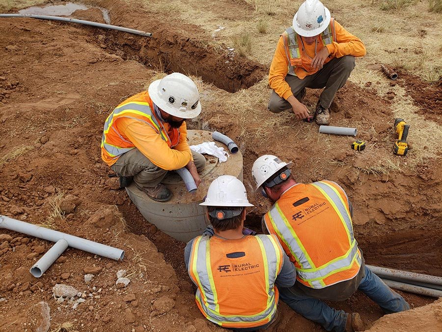 Four construction workers in orange safety vests and helmets install gray pipes into a concrete structure in a dirt trench for a Home - Rural Electric project.