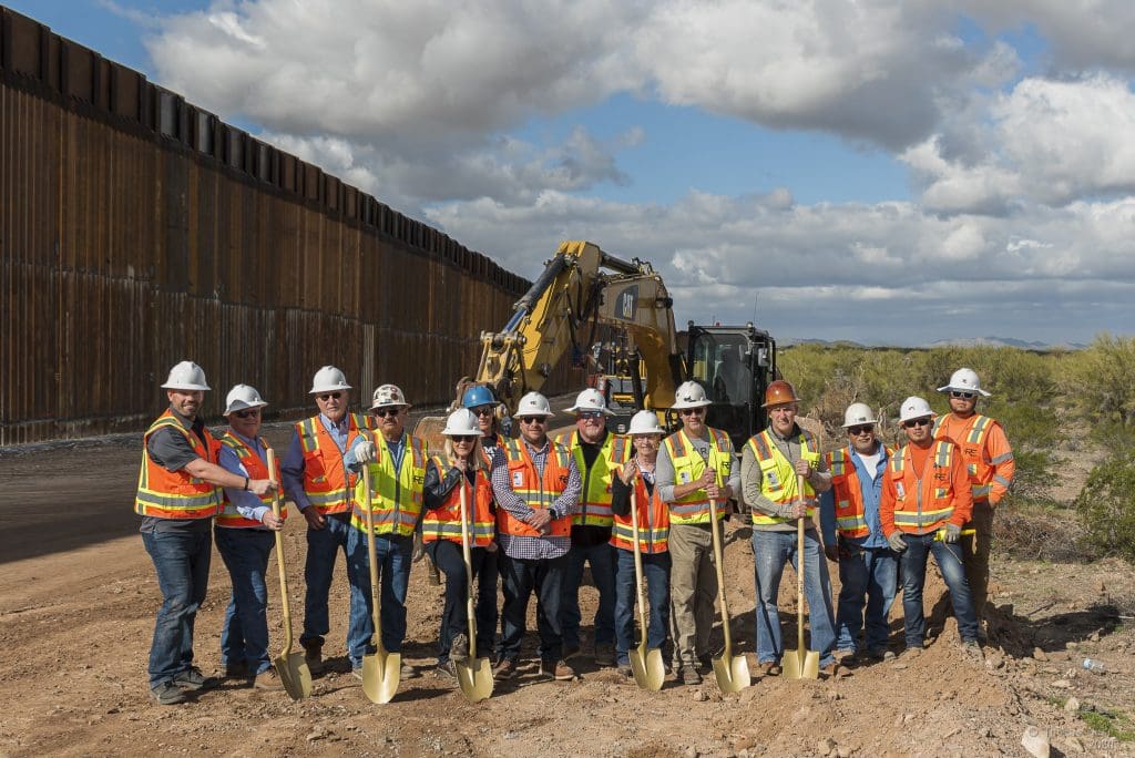 A group of construction workers in safety vests and helmets pose with shovels in front of heavy machinery and a border wall at a Home - Rural Electric dirt construction site.