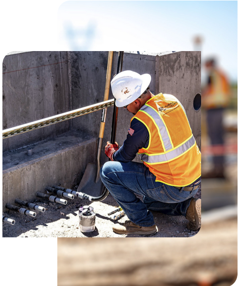 A construction worker in a hard hat and safety vest kneels by a concrete structure, handling tools and pipes at a Home - Rural Electric outdoor worksite.