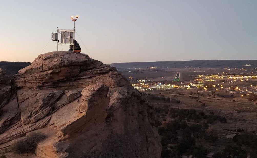 A weather monitoring station sits atop a rocky cliff at dusk, overlooking a distant town with illuminated buildings and streets powered by rural electric homes.