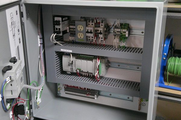 Open electrical control panel showing wiring, relays, outlets, terminal blocks, and other components—typical in Home – Rural Electric setups—with spools of green and white wire visible in the background. Open electrical control panel showing wiring, relays, outlets, terminal blocks, and other components—typical in Home - Rural Electric setups—with spools of green and white wire visible in the background.