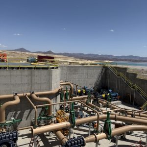 Industrial mining facility with large tan pipelines, machinery, and concrete structures outdoors under a clear blue sky, with distant mountains and water in the background. Industrial mining facility with large tan pipelines, machinery, and concrete structures outdoors under a clear blue sky, with distant mountains and water in the background.