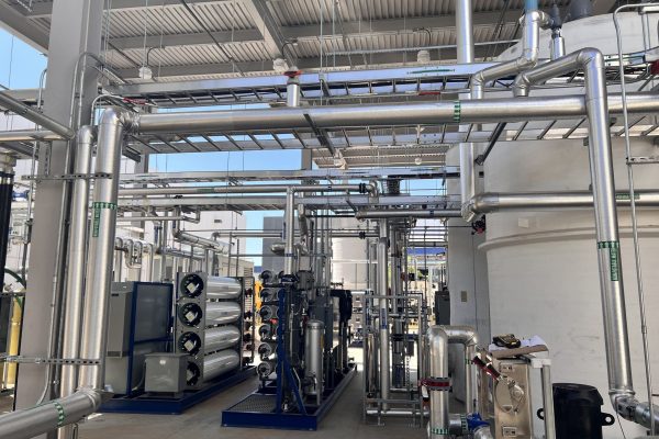 Industrial facility interior with interconnected stainless steel pipes, large white tanks, filtration units, and machinery under a metal roof with natural light, supported by rural electric infrastructure. Industrial facility interior with interconnected stainless steel pipes, large white tanks, filtration units, and machinery under a metal roof with natural light, supported by rural electric infrastructure.