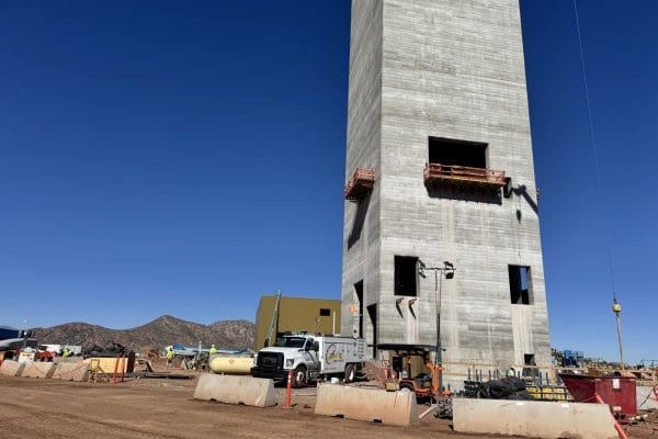 A tall concrete tower under construction stands on a dirt mining site with construction vehicles, equipment, barriers, and mountains in the background under a clear blue sky. A tall concrete tower under construction stands on a dirt mining site with construction vehicles, equipment, barriers, and mountains in the background under a clear blue sky.
