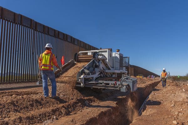 Construction workers operate heavy machinery to dig a trench alongside a tall metal border fence under a clear blue sky as part of Special Projects – Rural Electric initiatives. Construction workers operate heavy machinery to dig a trench alongside a tall metal border fence under a clear blue sky as part of Special Projects - Rural Electric initiatives.