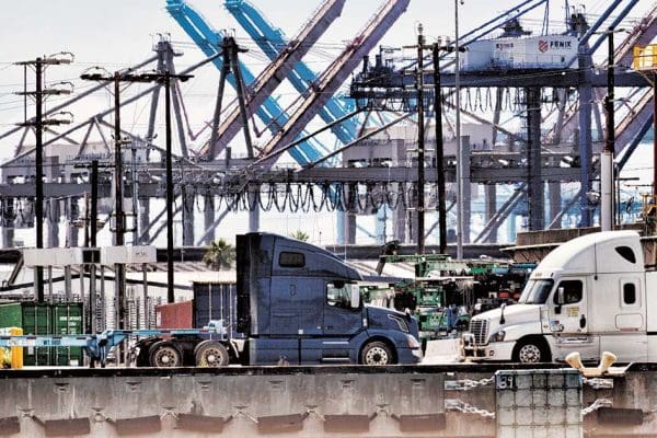 Two semi-trucks drive past shipping containers and cranes at an industrial port facility, highlighting the vital role of rural electric infrastructure in powering operations. Two semi-trucks drive past shipping containers and cranes at an industrial port facility, highlighting the vital role of rural electric infrastructure in powering operations.
