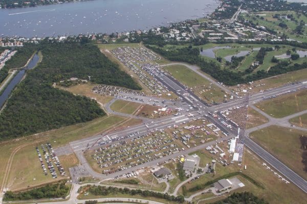 Aerial view of an airport with parked cars, buildings, and people gathered on the runway, near a waterfront and residential areas, showcases the close-knit community supported by rural electric infrastructure and local manufacturing. Aerial view of an airport with parked cars, buildings, and people gathered on the runway, near a waterfront and residential areas, showcases the close-knit community supported by rural electric infrastructure and local manufacturing.