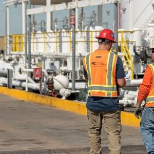 Two workers wearing safety vests and helmets walk and talk near industrial piping and equipment at an outdoor facility. Two workers wearing safety vests and helmets walk and talk near industrial piping and equipment at an outdoor facility.