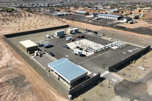 Aerial view of an industrial facility with various equipment, vehicles, and buildings, surrounded by a concrete wall in a desert area, possibly supporting rural electric infrastructure, with distant residential and industrial structures. Aerial view of an industrial facility with various equipment, vehicles, and buildings, surrounded by a concrete wall in a desert area, possibly supporting rural electric infrastructure, with distant residential and industrial structures.